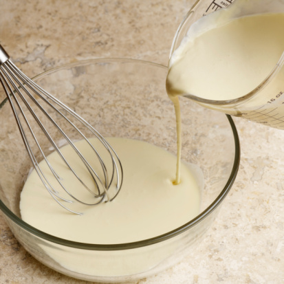 a glass jug pouring creamy yellow batter into a glass bowl and a silver metal whisk