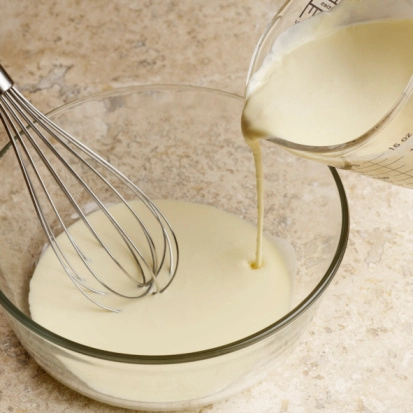 a glass jug pouring creamy yellow batter into a glass bowl and a silver metal whisk