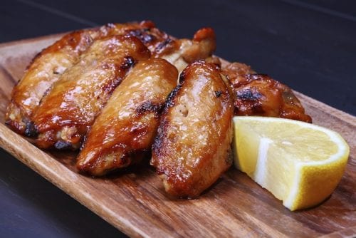 a brown wooden platter of deep brown, crispy cooked chicken wings and a slice of yellow lemon, liquid smoke chicken wings