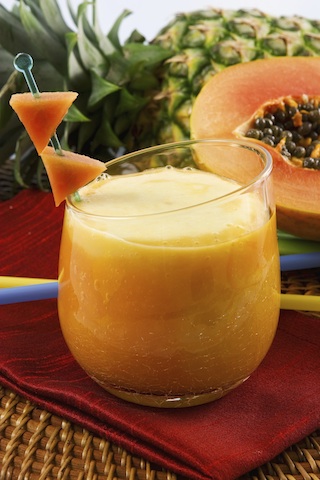 a glass of orange tropical fruit juice on a brown table against a backdrop of tropcial fruit, mango papaya and pineapple