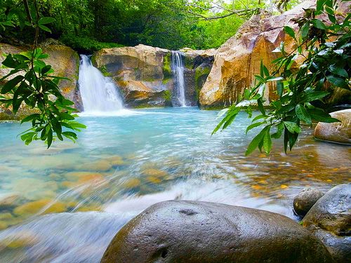 short waterfall over brown rocks into a blue pool surrounded by green foliage