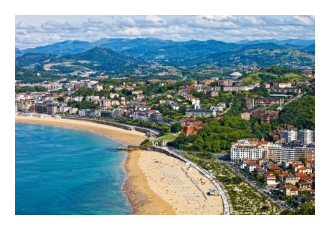 looking down onto a yellow beach, green hills, red roofed buildings against a blue sea and blue sky