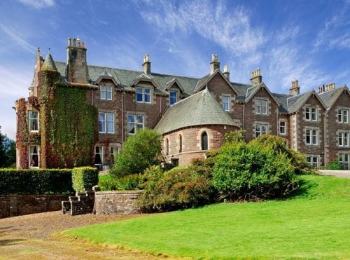 a large red brick Victorian hotel with white windows surrounded by green grass and green bushes against a blue sky