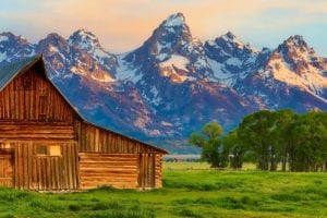 A brown wood cabin on green grass against a snowy mountain backdrop, Jackson Hole Wyoming