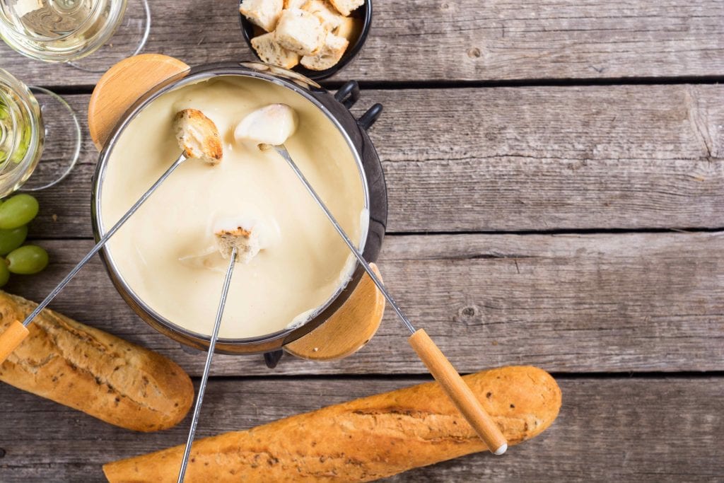 looking down onto a brown bowl with melted cheese and silver fondue sticks on a wooden table