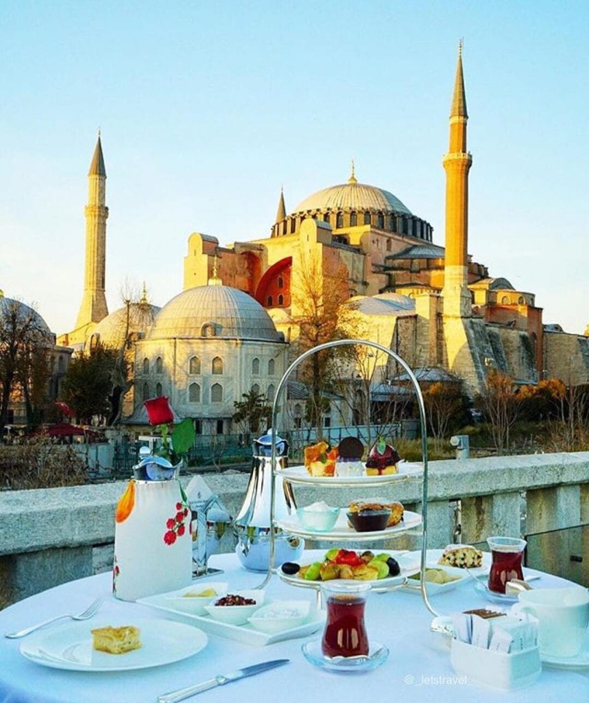 a white table set for an afternoon tea with cakes and sandwiches against the backdrop of a mosque and blue sky