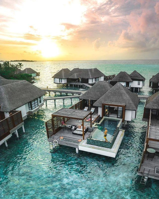 brown thatched huts on stilts over turquoise water against a sunset back drop