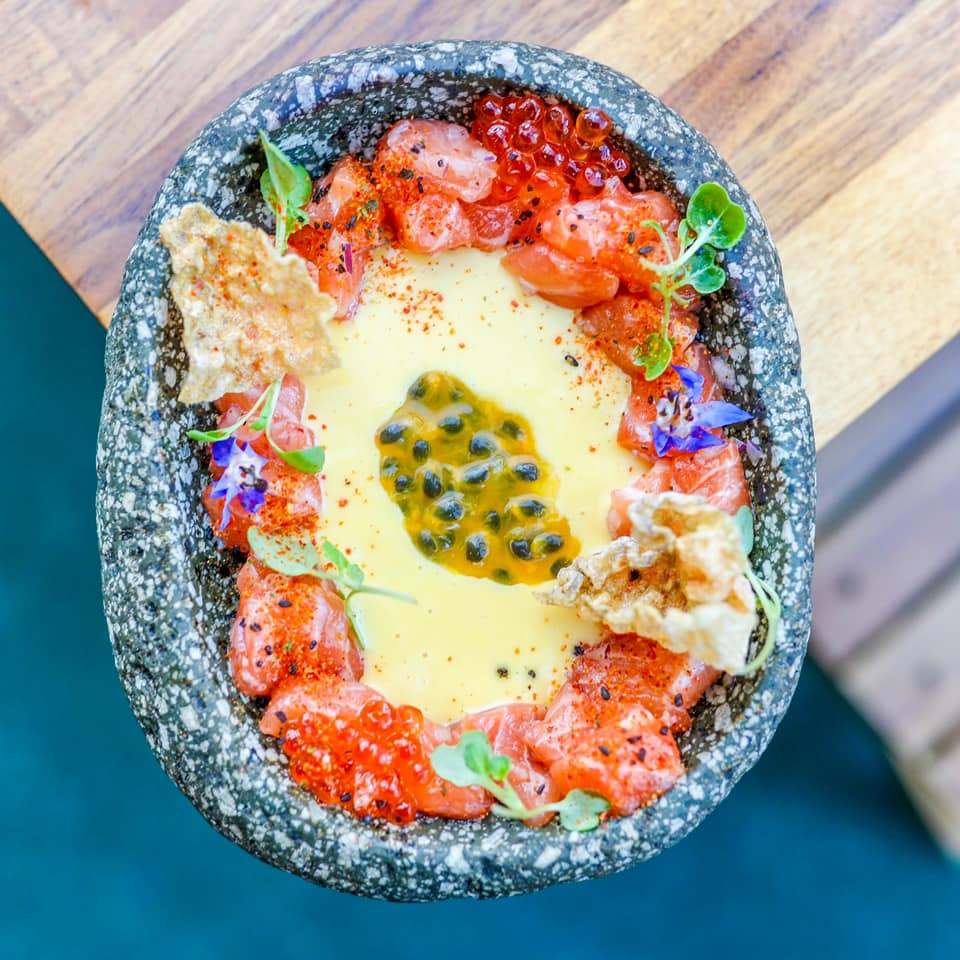 Looking down onto a blue stone bowl of food, red tomatoes around the edge with a yellow sauce and passionfruit seeds in the middle, ceviche nyc