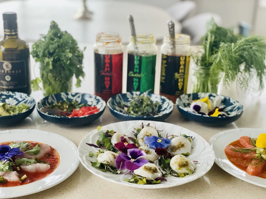 3 jars of sauce coloured red, green and yellow. glass jars of green leaves, 3 white plates of colourful food on a white table against a white background