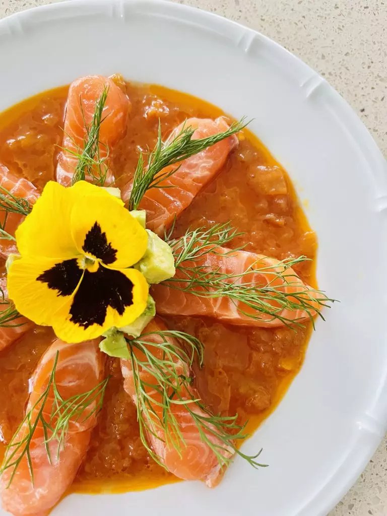 a white plate of food of orange sauce, raw orange pink salmon slices topped with a green leaf and a yellow flower