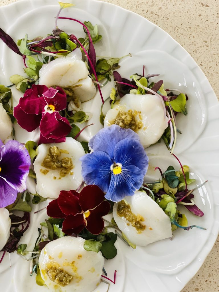 a white plate of food containing white raw fish, green sauce, green and purple salad leaves topped with purple and blue flowers