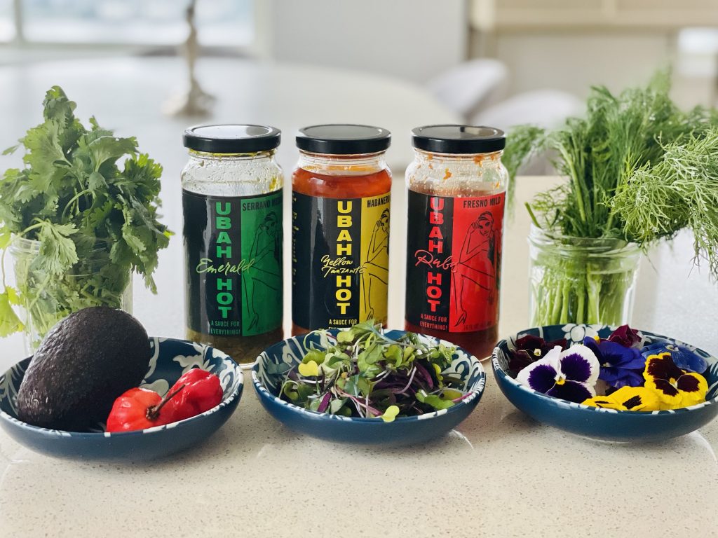 three jars of Ubah Hot Sauce behind three blue plates of vegetables and white and yellow flowers, two glass jars of green shoots and leaves, on a white table against a white background