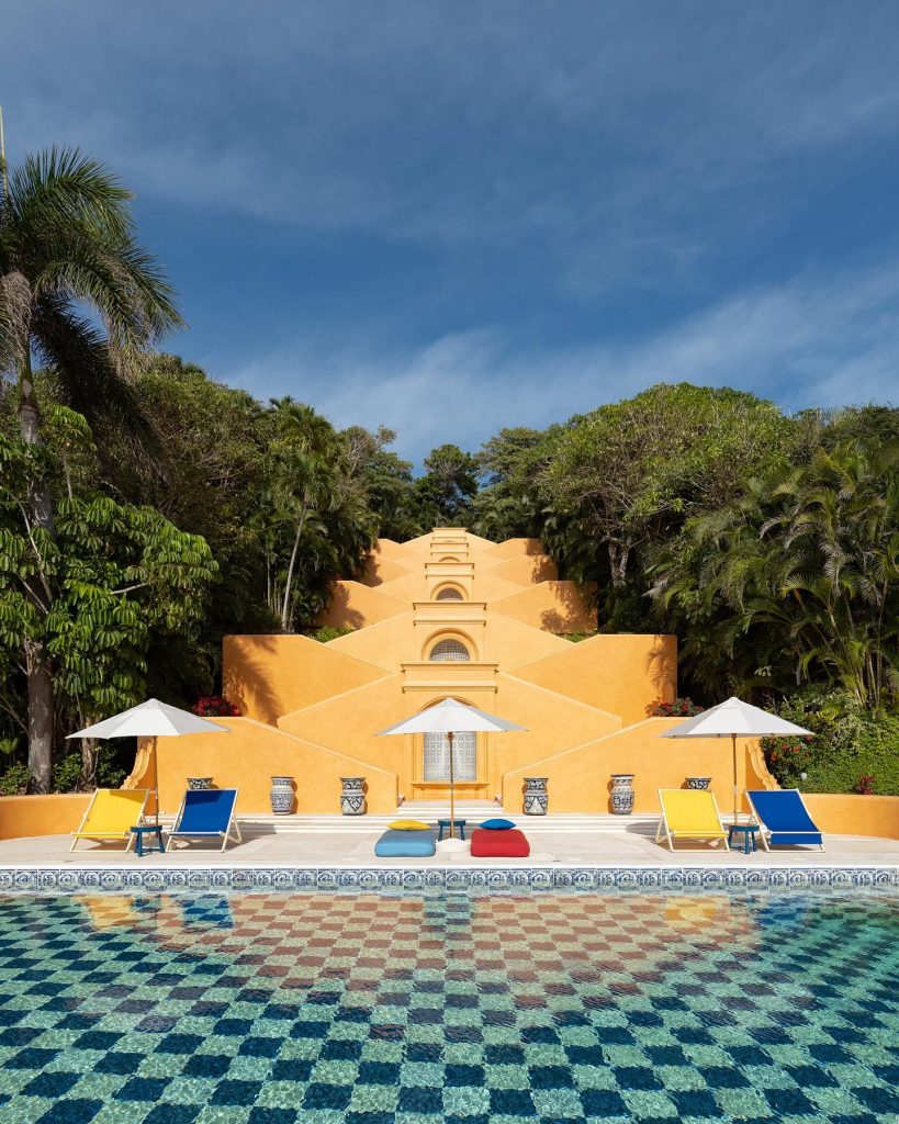 terracotta building in front of a swimming pool with a checked tile mosaic, backdrop of green trees and blue sky