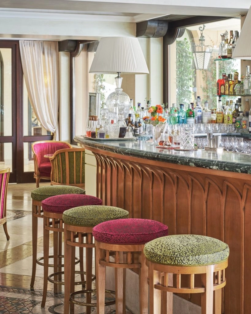red and green topped bar stools in front of a wood bar with marble top, covered in glasses and lamps