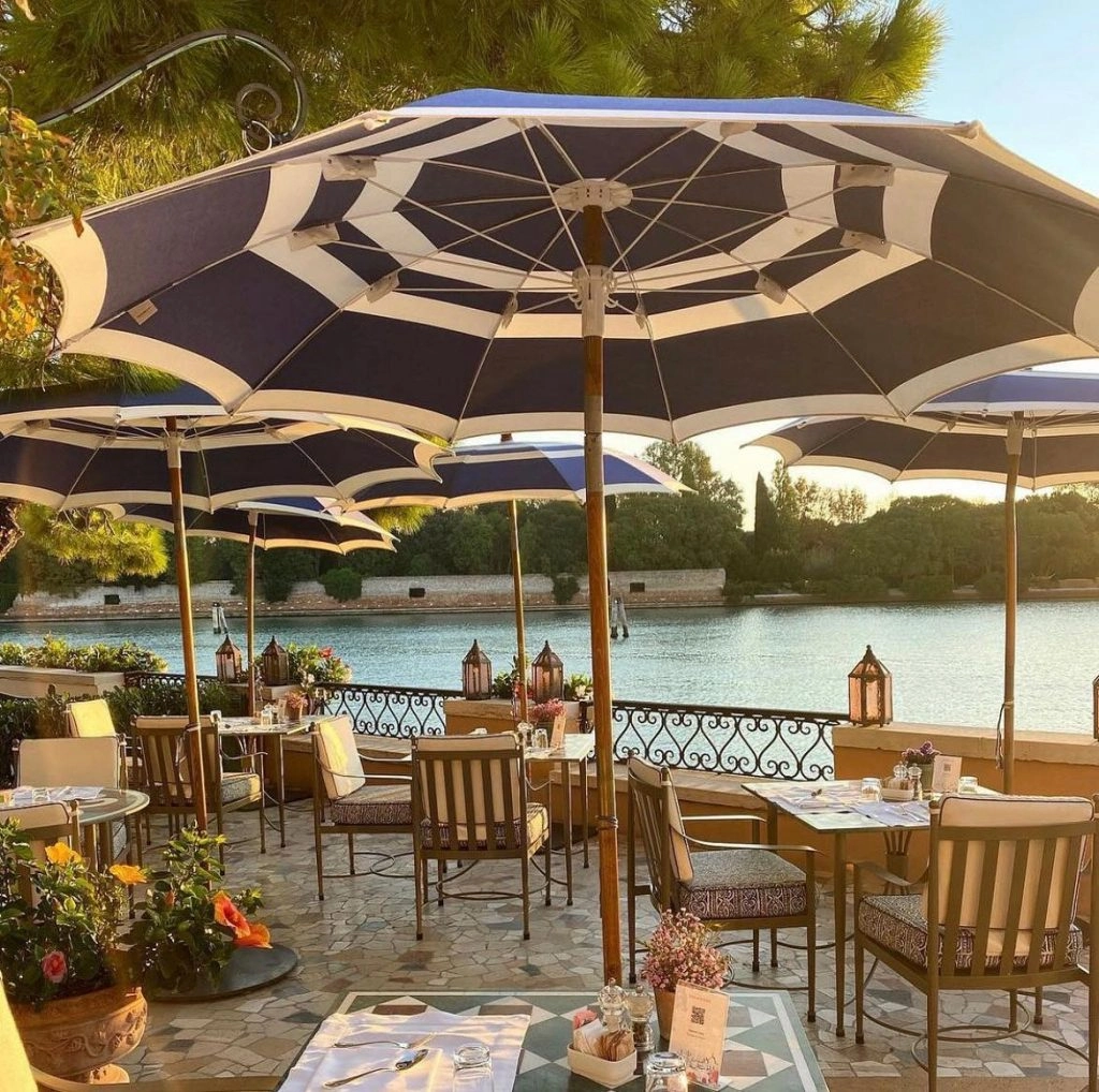 wooden tables and chairs with large blue and white sun umbrellas against a backdrop of water and green trees