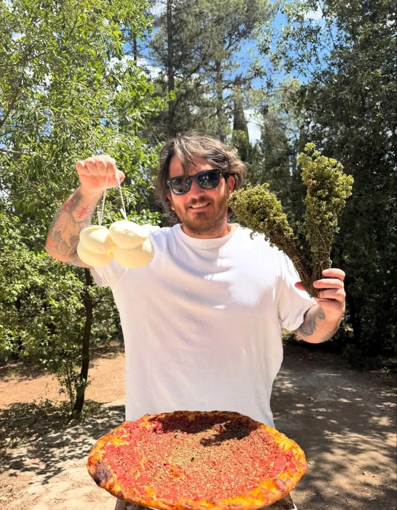 Chef holding fresh mozzarella and herbs above a rustic pizza outdoors in a forest setting at Baby Dicecca in Italy