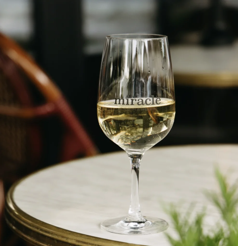 A glass of chilled white wine sits on a marble café table at Miracle restaurant in Sag Harbor, featuring the restaurant’s logo on the glass in a softly lit dining setting.