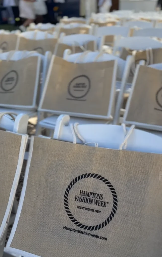 Rows of white event chairs at Hamptons Fashion Week featuring branded tote bags at an outdoor luxury lifestyle event.